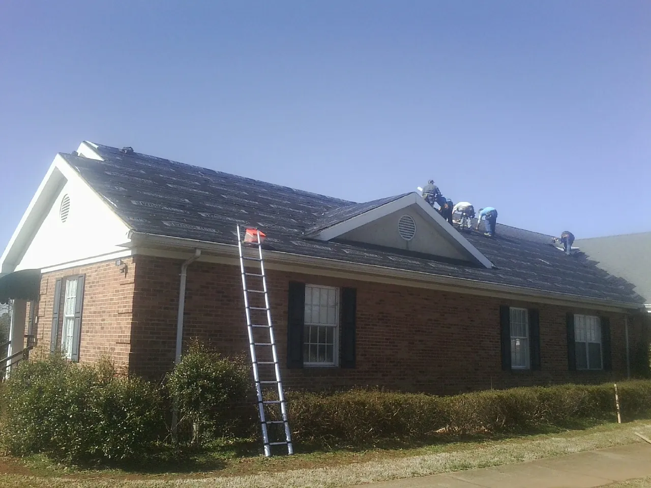 Crew tearing shingles off a brick ranch roof with a ladder set — Metal Roof Repair work by our crew in Triadelphia