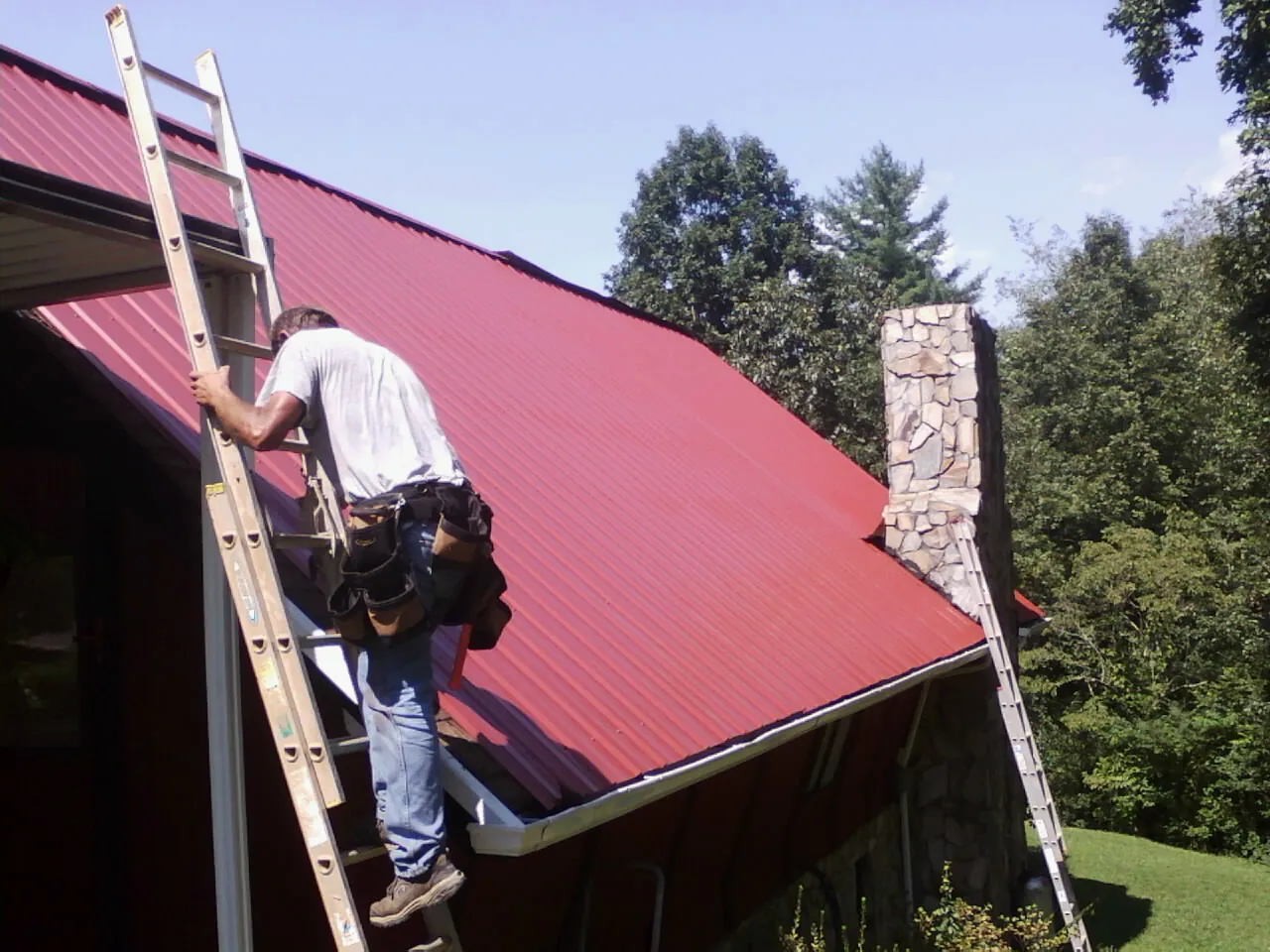 Roofer on a ladder beside a red metal roof and stone chimney — Soffit & Fascia Repair by Roofing Brothers in Triadelphia